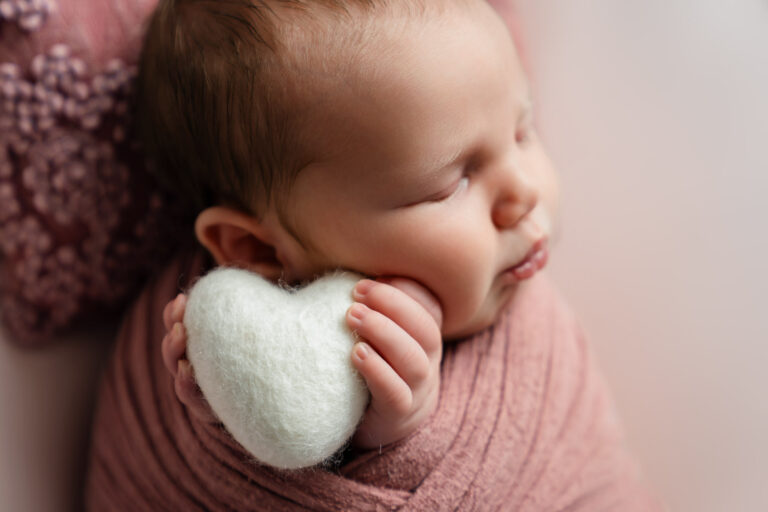 Newborn baby girl in a pink wrap, holding a white felt heart at a photoshoot near Newbury.