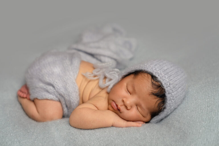 Newborn baby wearing a knitted pale grey hat and matching wrap, sleeping on a soft grey blanket at a photoshoot near Basingstoke.