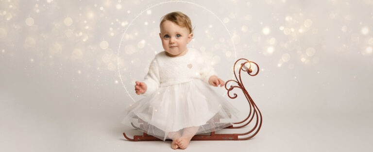 Baby girl sitting on a tiny sleigh, surrounded by Christmas sparkles.