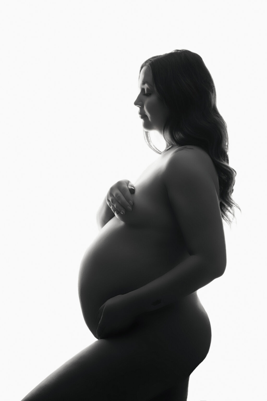 A nude pregnant woman cradles her bump, in a light and airy portrait by H Jones Photography in Andover, Hampshire.