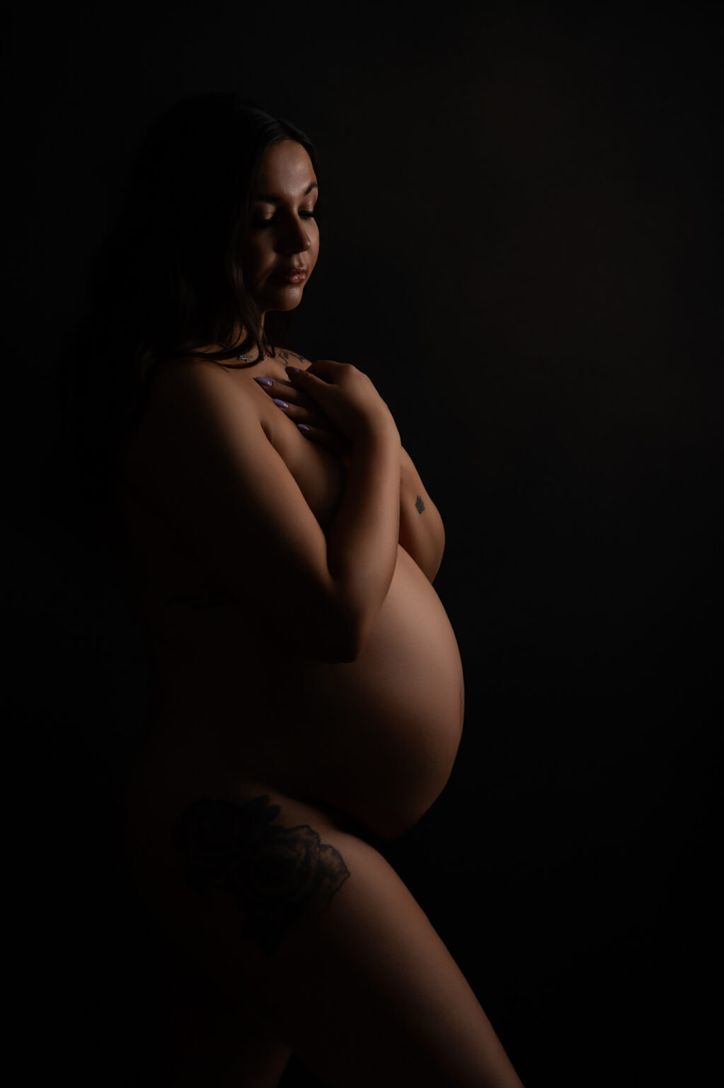 A pregnant woman in a dark, moody portrait cradles her bump, softly lit to highlight her serene expression. Captured in a professional pregnancy photoshoot in Andover, Hampshire.