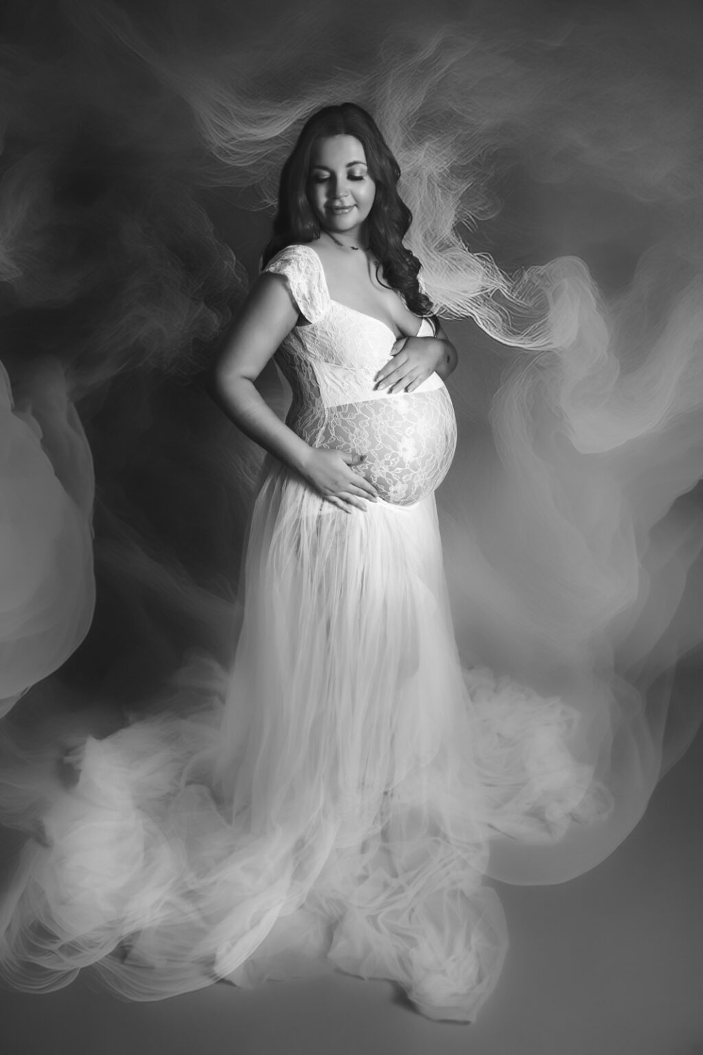 A woman in a white flowing dress, with lace front exposing her pregnant belly, poses standing in a mist of white smoke at a maternity photoshoot in Hampshire.