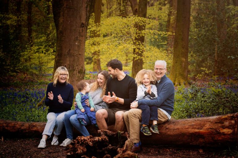 Family of four plus grandparents sitting on a fallen log and laughing naturally at a bluebell photoshoot in Hampshire