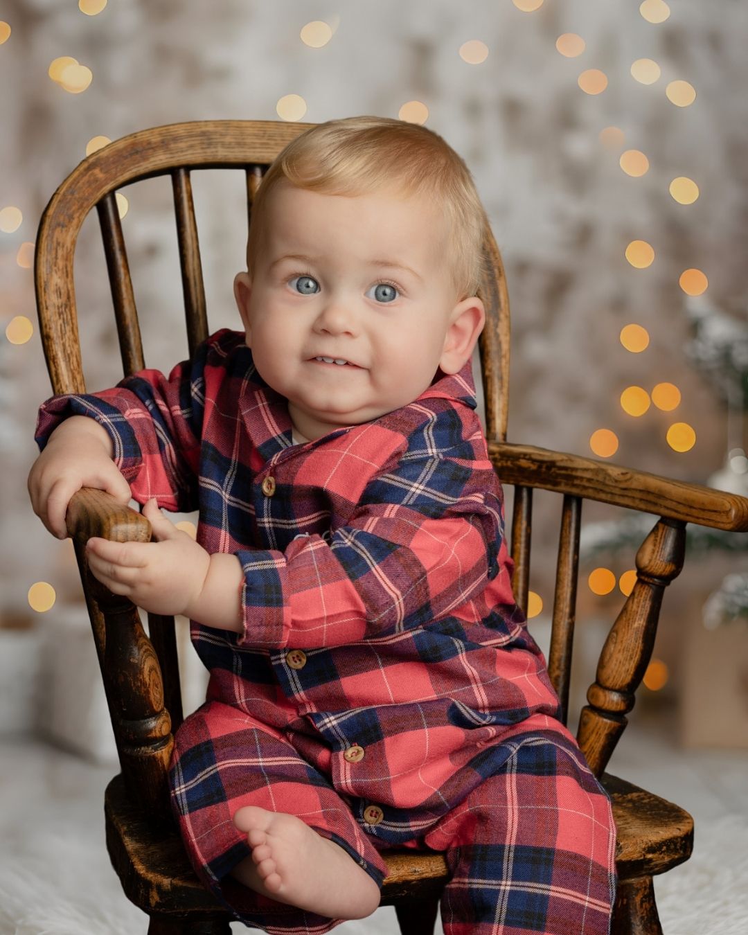 Baby boy in festive red and navy plaid pyjamas sitting on a wooden chair during a magical Christmas photoshoot in Hampshire.