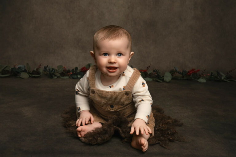 One year old boy in brown dungarees and gingerbread t shirt sitting on a small brown fur rug at Helen Jones photography studio.