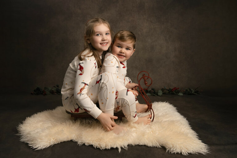 Brother and sister wearing white Christmas pyjamas, sitting on a small sleigh at H Jones Photography Christmas shoot.