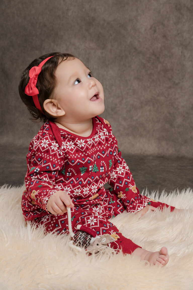 One year old girl in Christmas pyjamas wearing a red ribbon in her hair, sitting on a white fur rug at a photoshoot in Hampshire.