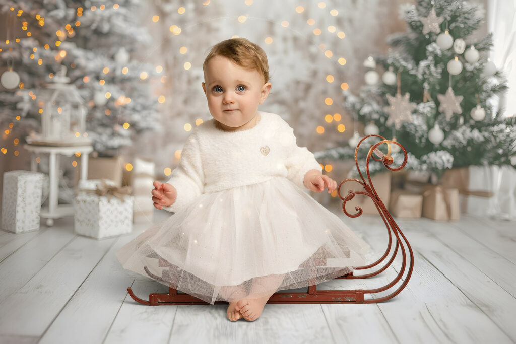 Baby sitting on a vintage red sleigh during a magical Christmas photoshoot in a cosy Hampshire studio, wearing a white festive dress with sparkling lights, Christmas tree and wrapped gifts in the background.