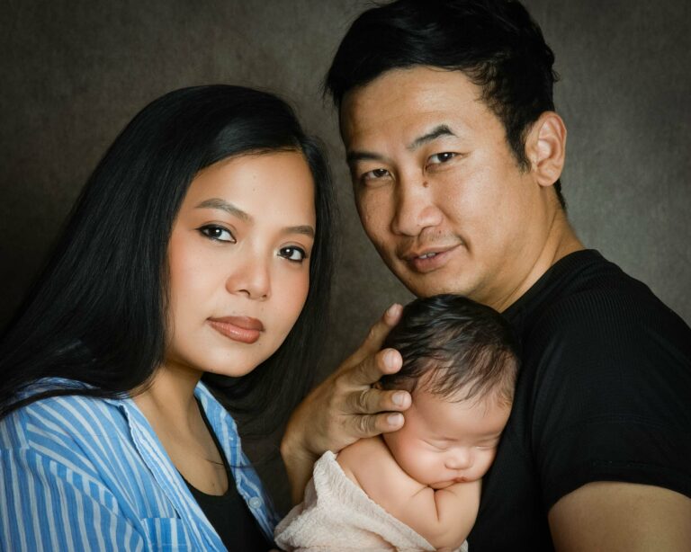 Portrait of a newborn baby sleeping peacefully against their father’s chest, with both parents holding their baby closely and looking towards the camera, captured in a warm and intimate family photoshoot in a professional Hampshire studio.