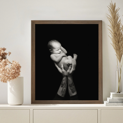Framed black-and-white newborn portrait displayed on a modern sideboard, showing a baby held safely in tattooed hands, styled as timeless wall art by a Hampshire newborn photographer.