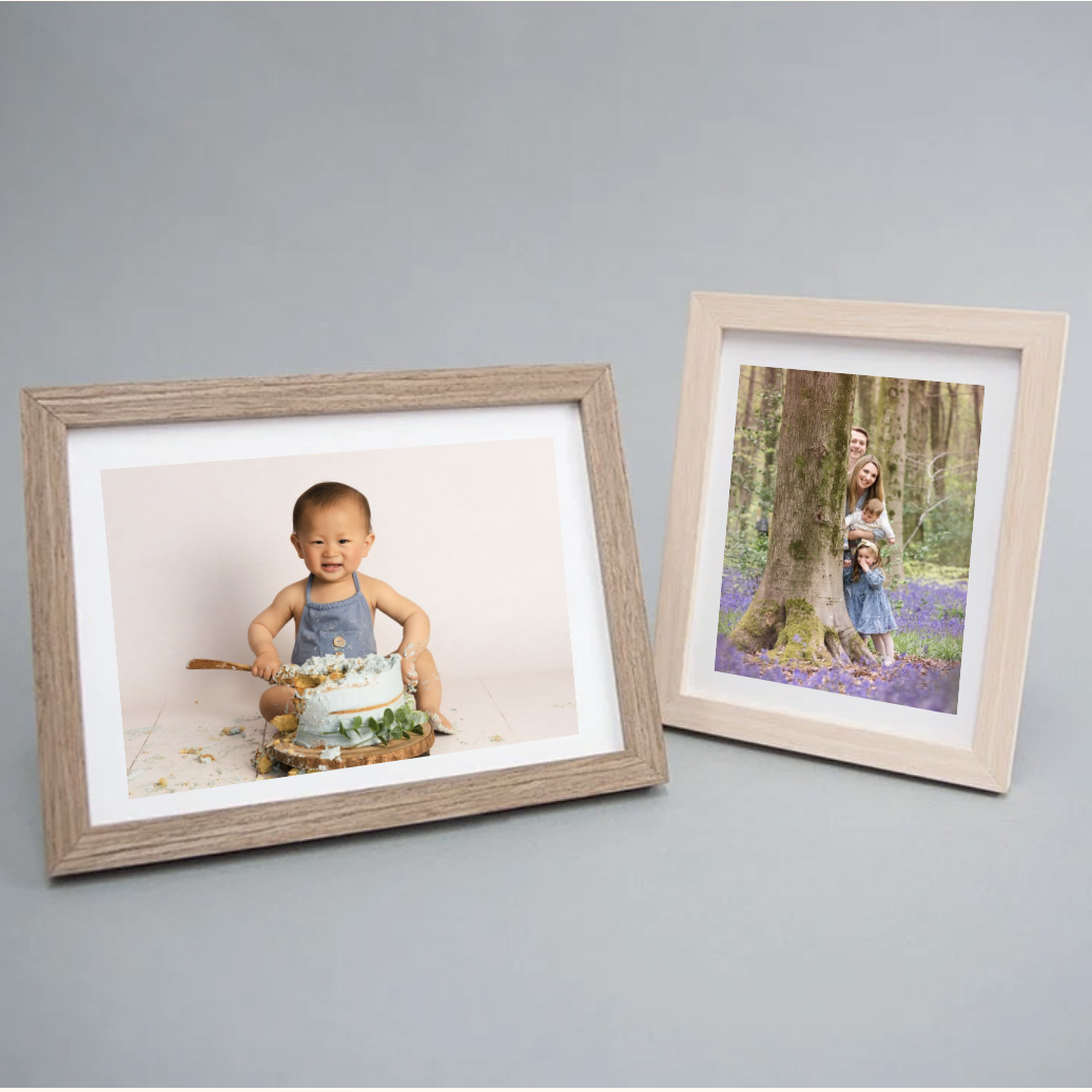 Two framed photography prints on a grey background: one featuring a smiling baby during a cake smash session, and the other showing a family standing among bluebells in a woodland setting.