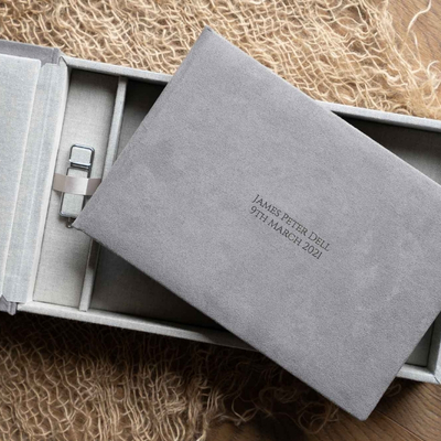 Soft grey suede photo album personalised with engraved text, presented inside a matching keepsake box with USB storage, styled on a textured natural fabric backdrop.
