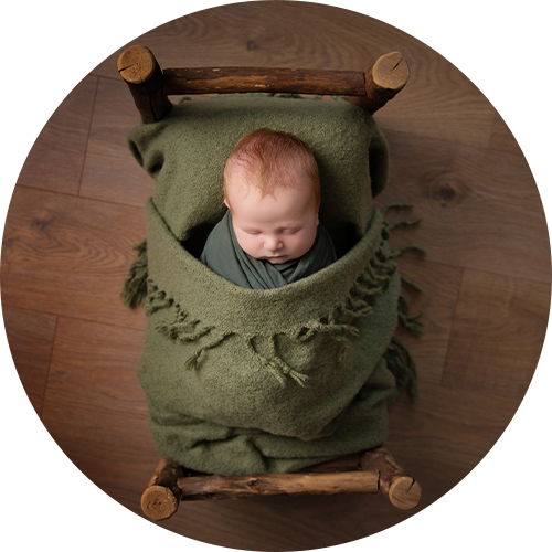Newborn baby peacefully swaddled in soft green blankets, lying in a rustic wooden prop bed on a warm wooden floor — a cosy, natural-style setup used in H Jones Photography testimonial sessions.