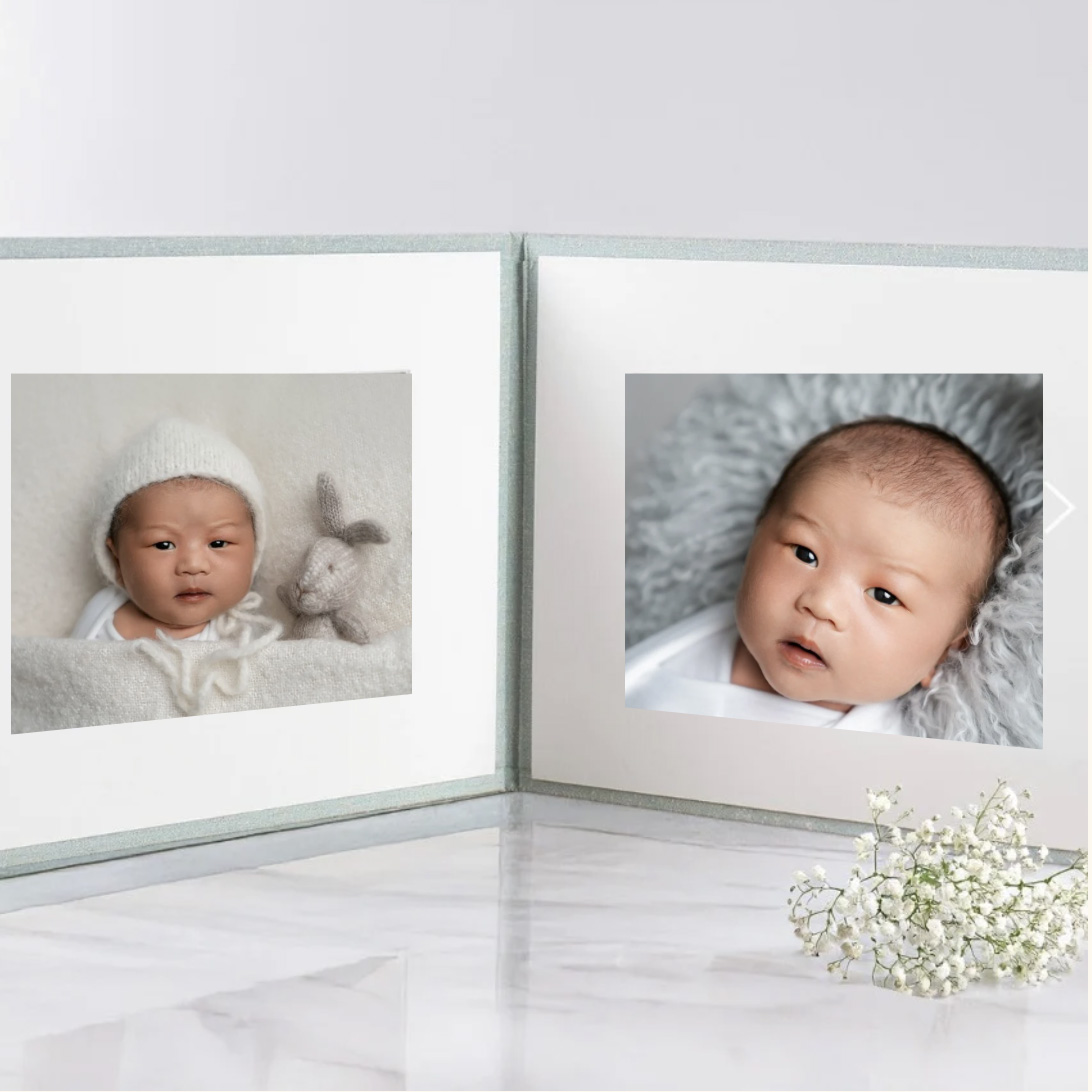 Beautiful newborn photography folio displayed open on a marble surface, featuring two soft, neutral-toned portraits of a baby—one snuggled in a white blanket with a knitted bonnet and tiny bunny, and the other resting on a fluffy grey backdrop—with delicate white flowers beside the album.