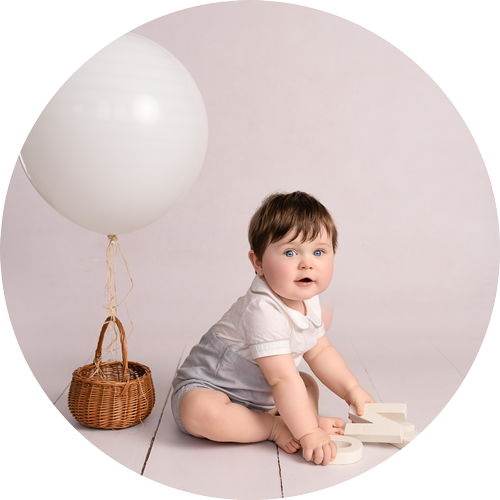 One-year-old baby sitting on a pale backdrop beside a white balloon, photographed during a first birthday cake smash photoshoot near Andover, Hampshire.
