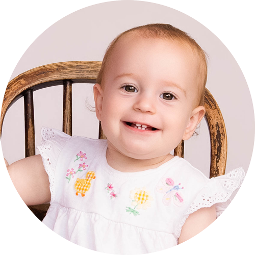 Smiling one-year-old girl sitting on a rustic wooden chair during her first birthday photoshoot at H Jones Photography's studio near Andover, Hampshire.