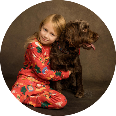 A young girl in red Christmas pajamas sits beside and hugs a large shaggy brown dog, both posed against a warm brown studio backdrop inside a circular frame.