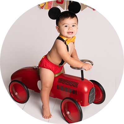 Smiling baby wearing black mouse ears, a yellow bow tie and red shorts, standing beside a vintage red toy car.