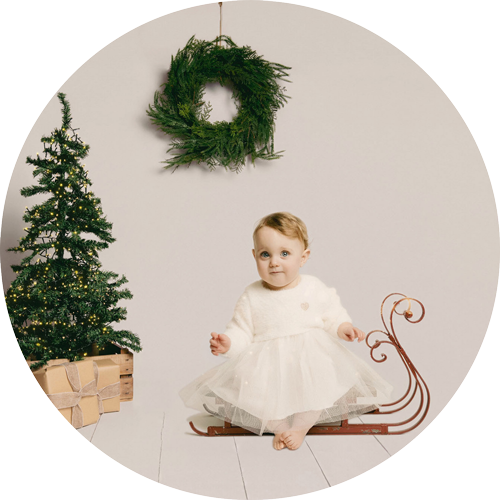 Child in a cream dress sitting on a vintage sleigh beside a Christmas tree with twinkling lights, wrapped gifts and a foliage wreath.